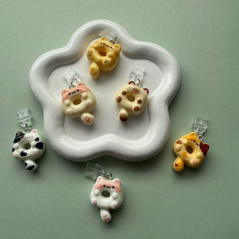 May include: A collection of six cat-shaped donut earrings in various colors, including yellow, pink, white, and black. The earrings are displayed on a white, cloud-shaped tray against a green background. Each earring has a silver-colored clip.