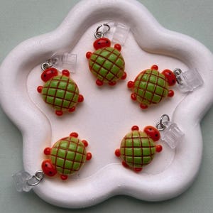May include: Five turtle-shaped charms. Each charm has a green shell with a red grid pattern, red head, and red feet. The charms are attached to clear plastic earring backs with silver-colored metal loops. The charms are arranged on a white, flower-shaped surface.