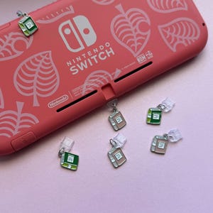 May include: A coral-colored Nintendo Switch console featuring a white leaf pattern and five small metal book-shaped charms. The charms are enamel-filled in shades of green and light pink, each attached to a clear plastic clip for attachment.