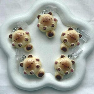 May include: Five cream-colored bear-shaped charms with brown and pink details, each attached to a silver-tone metal clasp. The charms are arranged on a white, flower-shaped tray. The bears have small, round ears and paws.