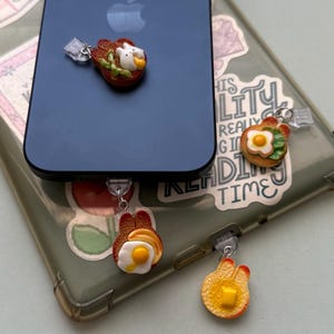 May include: A collection of food-themed phone charms. The charms include a piece of toast with a fried egg, a piece of toast with butter, and a piece of toast with a bird and vegetables. The charms are attached to a phone case.