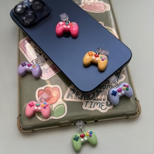 May include: A collection of colorful miniature game controller charms in pink, yellow, purple, light blue, and green. Each charm has a silver-colored clasp. The charms are attached to a phone case with a floral design.