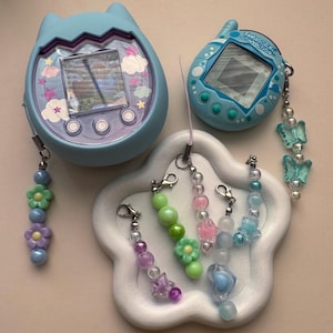May include: Two light blue Tamagotchi virtual pet devices with attached beaded charms. The charms feature various pastel-colored beads, flowers, and stars. One device is egg-shaped, the other is rectangular. The devices are on a white, cloud-shaped tray.