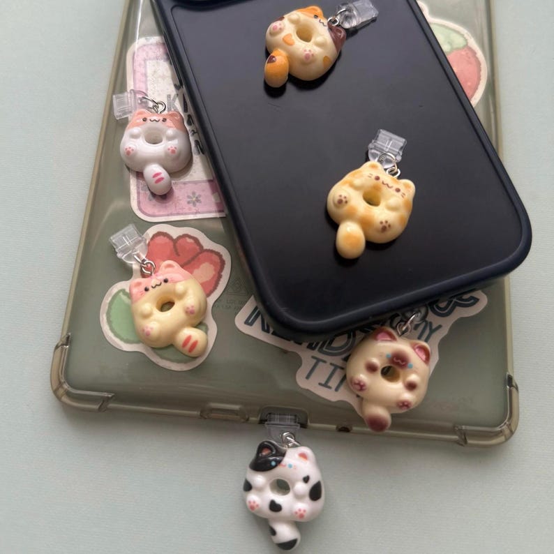 May include: A collection of cat-shaped phone charms in various colors, including white, orange, and black. The charms are attached to a black phone case and a clear phone case with stickers.