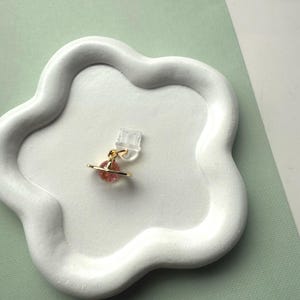 May include: A gold-colored earring with a pink planet-shaped charm and a clear plastic backing. The earring is displayed on a white, cloud-shaped tray. The background is a light green color.