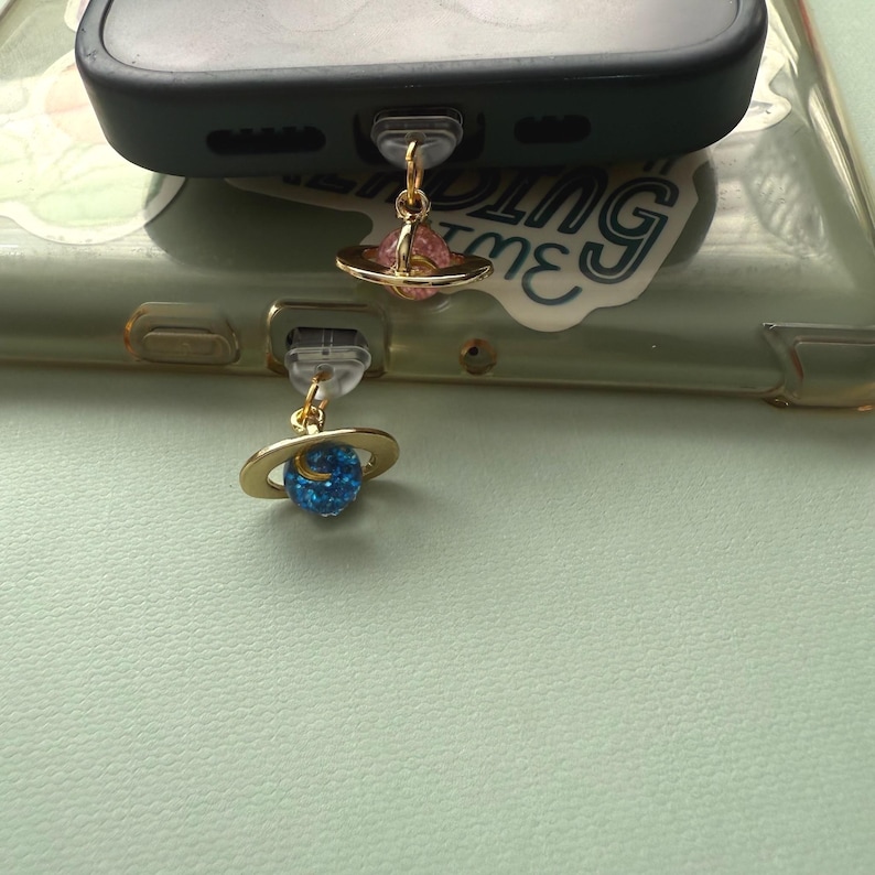 May include: Two gold-colored planet charms attached to a phone. One charm features a pink planet with a gold ring, the other a blue planet with a gold ring. The phone is black and has a clear case.