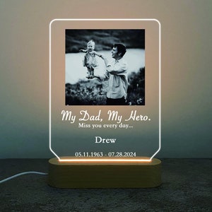 以下が含まれることがあります： 父親と娘の白黒写真がついた、パーソナライズされたアクリル製フォトランプ。ランプは木製ベースで、暖色系の白い光を放っています。ランプのテキストは「My Dad, My Hero. Miss you every day... Drew 05.11.1963 - 07.28.2024」です