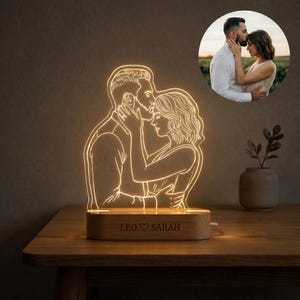 May include: A custom LED night light featuring a glowing line-art illustration of a couple embracing, based on the circular photo inset. The light is mounted on a rectangular wooden base engraved with the text LEO ♡ SARAH in a serif font.