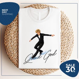 May include: White t-shirt featuring a graphic of a figure skater in a black outfit with the text "Quad God" in black script. The skater has blonde hair and is in a dynamic pose. The shirt is displayed on a woven surface.