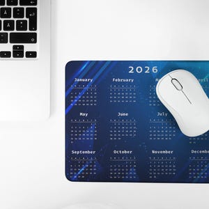May include: A blue mousepad with a 2026 calendar design and a white wireless mouse. The calendar features a dark blue background with a celestial design and the months of the year. A laptop keyboard is visible in the upper left corner.