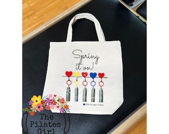 May include: White canvas tote bag with the text "Spring it on!" and a graphic of five colorful hearts with silver Pilates rings hanging from them. The text "PDX Flower Power" is below the graphic.