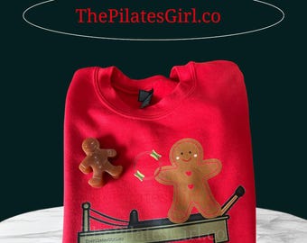 May include: Red sweatshirt with a gingerbread man design. The design includes a gingerbread man on a Pilates reformer, a smaller gingerbread man, and the text "ThePilatesGirl.co". The sweatshirt is a festive holiday item.