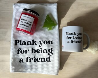 May include: A white waffle-weave towel, a red candle, a green tree-shaped ornament, and a white mug with the phrase "Plank you for being a friend" printed in black. The candle has a black lid.