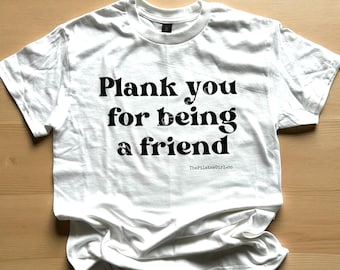 May include: White t-shirt with the phrase "Plank you for being a friend" in black lettering. The PilatesGirl.co logo is printed at the top and bottom of the shirt. The shirt is laying on a wooden surface.