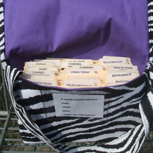 May include: A black and white zebra-print tote bag with a purple lining. The bag is open and filled with index cards labeled with different grocery categories, such as "Baking", "Dairy", "Canned", "Cleaning", "Rolls/Biscuits", "Health Food", "Restaurants", "Bags/Frozen", and "Salad Dressing". A small white card with the text "IF FOUND PLEASE CONTACT" is attached to the front of the bag.