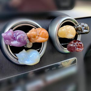 May include: Six colorful sleeping cat car air freshener charms. The charms are made of polished stone and are in the shapes of cats sleeping. The charms are hanging from the air vents of a car.