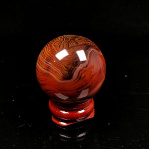 May include: A red agate sphere with brown and white banding, polished to a smooth finish. The sphere is displayed on a red wooden stand.