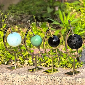 Puede incluir: Cuatro esculturas esféricas decorativas en fila. Cada esfera está montada sobre un soporte de metal dorado. Las esferas son azul claro, verde, negra y negra con superficie texturizada. Los soportes tienen un diseño curvo.