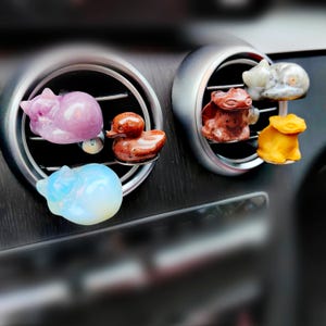 May include: Six small, colorful, animal-shaped car air fresheners. The fresheners are made of stone and are attached to the air vents of a car. The fresheners include a cat, a duck, a dragon, a frog, and two more cats.