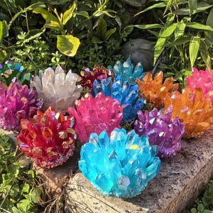 Puede incluir: Una colección de racimos de cristales coloridos en tonos rojos, rosas, naranjas, azules, morados y blancos. Los cristales están dispuestos sobre una superficie de piedra en medio de follaje verde, mostrando sus formaciones naturales y colores vibrantes.