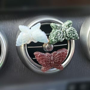 Puede incluir: Tres ambientadores de coche con forma de mariposa. Uno es blanco con detalles iridiscentes, otro es verde con motas blancas y el último es de color burdeos oscuro.