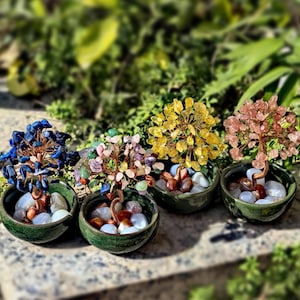 Könnte beinhalten: Vier dekorative Edelsteinbäume in kleinen grünen Keramiktöpfen. Die Bäume haben blaue, mehrfarbige, gelbe und rosa Edelsteine mit Kupferdrahtzweigen. Die Töpfe sind mit weißen und braunen Steinen gefüllt, vor einem verschwommenen grünen Hintergrund.