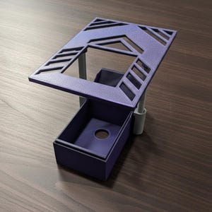 May include: A purple 3D printed desk accessory featuring a rectangular base with a central circular cutout and an elevated, angled top platform with geometric cutouts, supported by two light grey vertical pillars attached to the rear side.