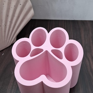 Puede incluir: Un organizador de escritorio rosa en forma de pata con múltiples compartimentos. El organizador está hecho de un material sólido y tiene una superficie texturizada. El diseño se asemeja a una huella de pata de mascota, con compartimentos redondeados. Hay plantas decorativas en el fondo.