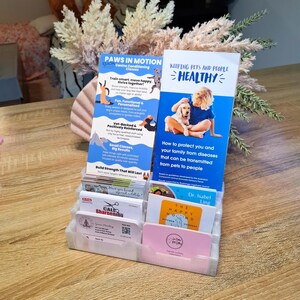May include: A clear plastic business card holder filled with various cards and brochures. The brochures advertise "Paws in Motion" canine conditioning classes and "Keeping Pets and People Healthy." Other cards include "Callo Sharpening" and "The Happy Hound."