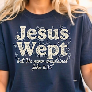 May include: Navy blue t-shirt with the text "Jesus Wept" in a decorative font, along with the quote "but He never complained John 11:35." The text is in a cream color with floral accents. The shirt is a casual wear item.
