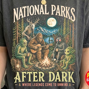 Könnte beinhalten: Ein anthrazitfarbenes Kurzarm-T-Shirt mit einem Grafikdruck und dem Text NATIONAL PARKS AFTER DARK WHERE LEGENDS COME TO UNWIND. Die Illustration zeigt eine Lagerfeuerszene mit einem Sasquatch, einem Mothman und weiteren Kreaturen.