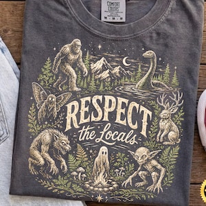 Puede incluir: Una camiseta gris carbón con un estampado gráfico que incluye el texto "RESPECT the Locals" rodeado de ilustraciones de Bigfoot, el monstruo del lago Ness, un hombre polilla, un jackalope, un hombre lobo, un fantasma y un duende.