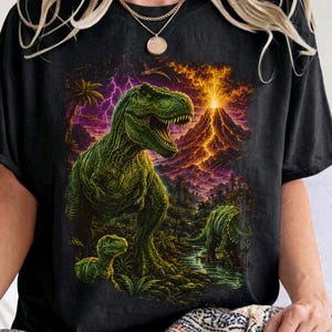 Op de afbeelding: Een zwart T-shirt met korte mouwen en een grafische print van een grote groene Tyrannosaurus Rex in een prehistorisch landschap met een vulkaanuitbarsting, paarse bliksem en een kleinere dinosaurus op de voorgrond.