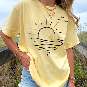 May include: A pale yellow short-sleeve t-shirt featuring a black line art graphic of a rising sun over wavy water with three small birds flying above. The shirt is paired with light blue denim shorts and layered gold necklaces and bracelets.