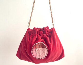 Puede incluir: Un bolso de terciopelo rojo vibrante con cierre de cordón, parte superior fruncida y una correa larga de cuerda beige trenzada. En el centro hay un parche circular hecho de retazos de tela cosidos en tonos rosa, marrón y crema.
