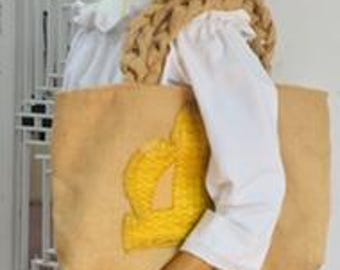 May include: A tan tote bag with a yellow emblem, carried by a mannequin. The bag has a rectangular shape and a simple design. A patterned scarf is draped over the mannequin's shoulder. The mannequin is wearing a white shirt and brown pants.
