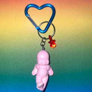 May include: A pink baby doll keychain with a blue heart-shaped carabiner clip and a red pacifier charm. The baby doll has a detailed face and is attached to a silver keyring.