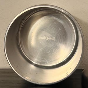 May include: A stainless steel bowl with a brushed finish. The bowl is round with a slightly flared rim. The interior of the bowl has a smooth, reflective surface. The bowl is empty and sits on a dark surface.