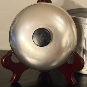 May include: A silver-coloured, spherical object with a black circular centre, resting on a dark red wooden stand. The stand has a decorative, curved design. The object appears to be a bell or similar decorative item.