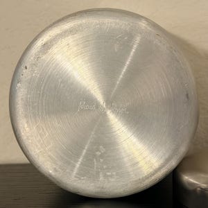 May include: The base of a silver-coloured pot with the words "Maid of Honor" etched into the metal. The pot has a circular design with concentric rings.
