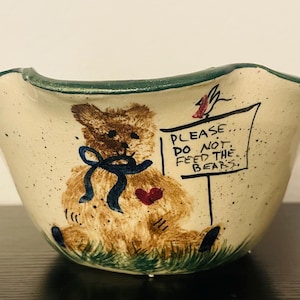 May include: A ceramic bowl with a cream-colored exterior and a green rim. The bowl features a hand-painted teddy bear with a blue bow and a red heart. Text on the bowl reads "PLEASE DO NOT FEED THE BEARS."