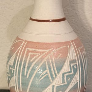 May include: A white ceramic vase with a rounded body and a narrow neck. The vase features a geometric design in pink and blue, with white carved lines. Two brown bands encircle the neck. The vase is a decorative home decor item.