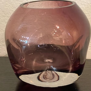Puede incluir: Un jarrón redondo de cristal transparente de color morado oscuro. El jarrón tiene una forma bulbosa con una abertura ancha y una base transparente y redondeada. El cristal refleja la luz, creando reflejos y sombras.