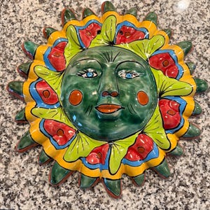 Puede incluir: Colgante de pared de cerámica con cara de sol verde, ojos azules y mejillas naranjas. Los rayos del sol son una mezcla de amarillo, verde, rojo y azul. Esta pieza decorativa es adecuada para uso en interiores o exteriores.