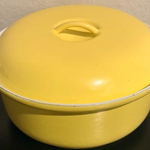 May include: A bright yellow enameled cast iron Dutch oven with a matching lid. The lid has a loop handle and the pot has white handles on either side. The pot is sitting on a dark surface.