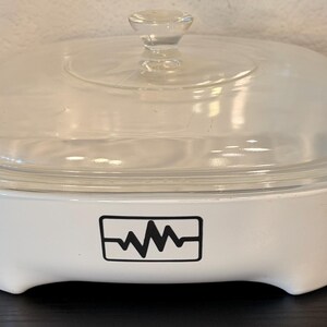 May include: A white rectangular ceramic casserole dish with integrated side handles and a clear glass lid. The front of the dish features a black rectangular graphic containing a stylized heartbeat line pattern centered on the exterior surface.