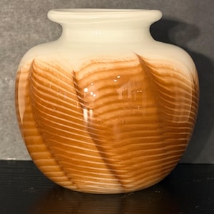 Può includere: Un vaso decorativo con corpo arrotondato e bordo svasato. Il vaso presenta una parte superiore color crema e una sezione inferiore con un motivo a vortice di tonalità marroni e crema. Il vaso di vetro è adatto per esporre fiori o come elemento decorativo.