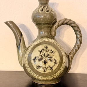 May include: An olive green ceramic teapot with a bulbous body, curved spout, and twisted handle. The teapot features a central floral design within a circular border. The top has a decorative finial. The design is reminiscent of traditional pottery.