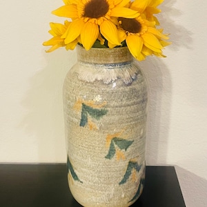 May include: A ceramic vase with a textured, speckled finish, adorned with green and yellow abstract shapes. The vase holds several bright yellow sunflowers with brown centers. The vase is on a black surface.