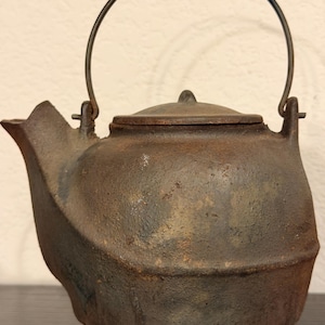 May include: A dark brown, textured cast iron teapot with a rounded body, a short spout, and a matching lid. The teapot features a thin, arched metal handle attached to two small vertical lugs on the top rim, showcasing a weathered, rustic finish.